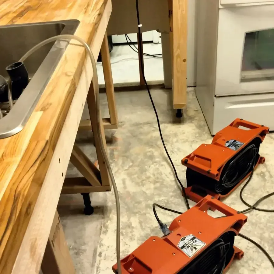 Orange air movers sit on stained flooring beside kitchen cabinets