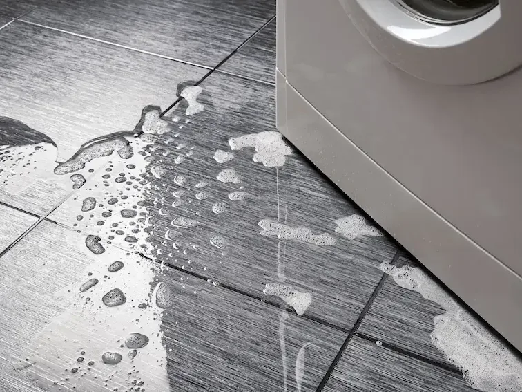 Water spreads across plank flooring beside the base of a washing machine