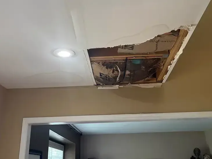 An opening in ceiling drywall exposes framing and pipes above the room