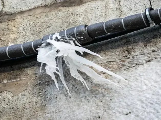 Ice hangs from a split black pipe running beside an exterior masonry wall.