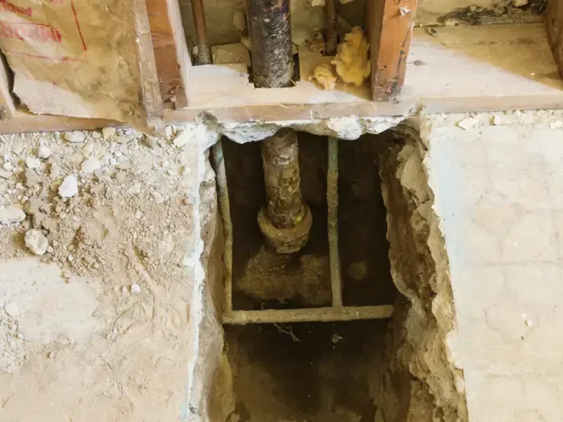 A cutout in a concrete floor exposes a pipe beneath an opened wall cavity